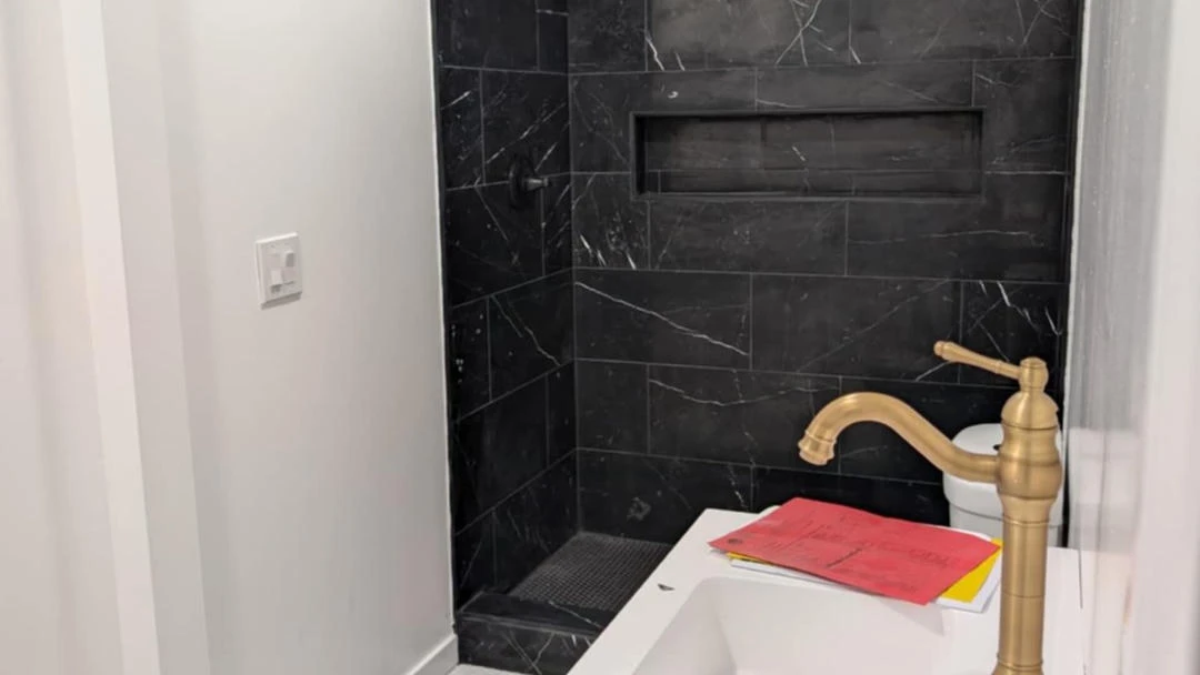 A bathroom with white walls and a shower tiled in black marble with white veins, featuring a brass faucet.